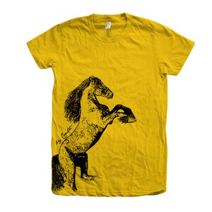 Op de afbeelding: Een geel T-shirt met een zwart-witte illustratie van een paard dat op zijn achterpoten staat. De tekst "Big Coat" is in cursief geschreven aan de linkerkant van de afbeelding.