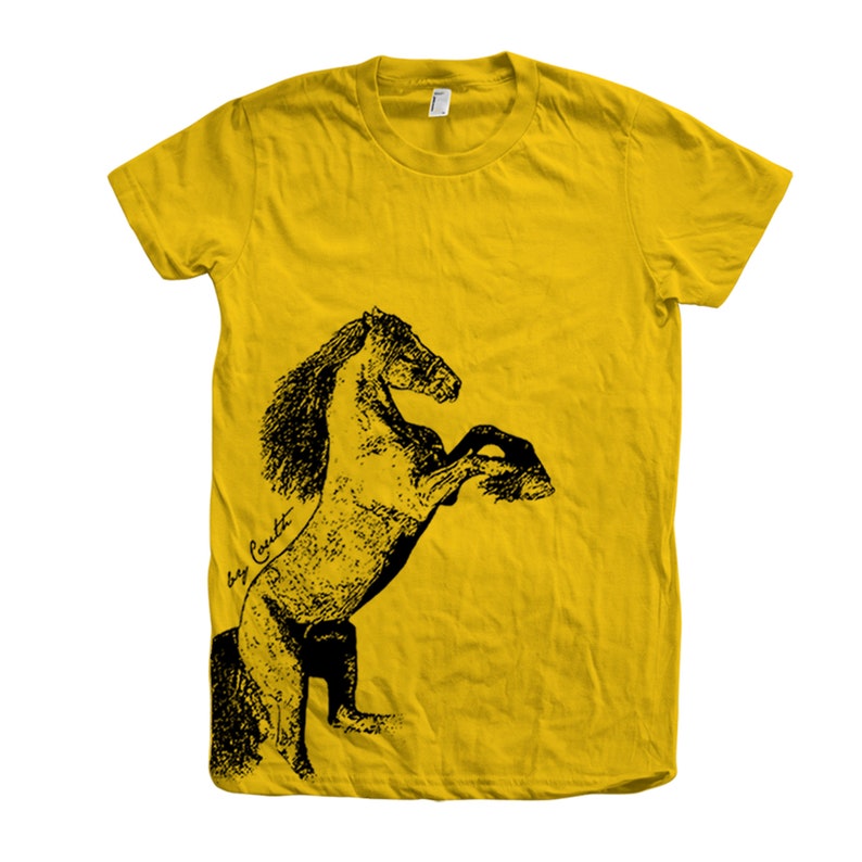Op de afbeelding: Een geel T-shirt met een zwart-witte illustratie van een paard dat op zijn achterpoten staat. De tekst "Big Coat" is in cursief geschreven aan de linkerkant van de afbeelding.