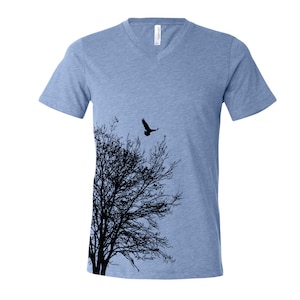 Könnte beinhalten: Hellblaues V-Ausschnitt-T-Shirt mit einem schwarzen Baum- und Vogelmotiv. Der Baum befindet sich auf der linken Seite, und der Vogel fliegt im Himmel. Das Shirt besteht aus einem weichen Material.