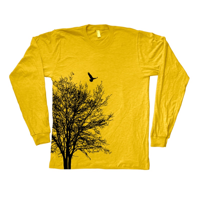 May include: A yellow long-sleeved t-shirt featuring a black silhouette of a tree and a bird flying above it. The shirt has a distressed look and the design is on the left side of the shirt.