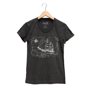 Puede incluir: Una camiseta gris oscuro con un gr&aacute;fico blanco de un barco navegando en un mapa con una br&uacute;jula. El texto "Coast" est&aacute; impreso en la camiseta.