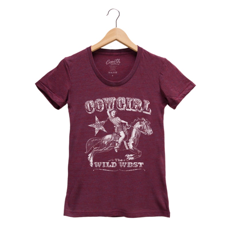 K&ouml;nnte beinhalten: Burgunderfarbenes T-Shirt mit einem wei&szlig;en Aufdruck einer Cowgirl, die ein Pferd reitet. Der Aufdruck enth&auml;lt den Text "COWGIRL" und "The WILD WEST".