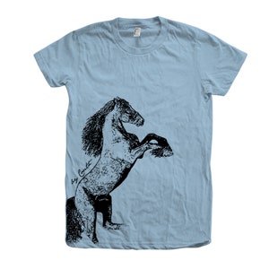 Op de afbeelding: Een lichtblauw T-shirt met een zwart-witte illustratie van een paard dat op zijn achterpoten staat. De illustratie is gesigneerd met "Bay Crockett".