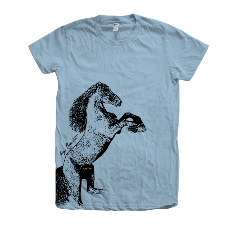 Op de afbeelding: Een lichtblauw T-shirt met een zwart-witte illustratie van een paard dat op zijn achterpoten staat. De illustratie is gesigneerd met "Bay Crockett".