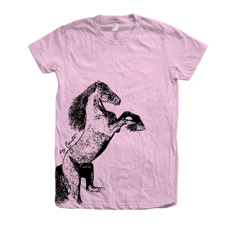 Op de afbeelding: Een roze T-shirt met een zwart-witte illustratie van een paard dat op zijn achterpoten staat. De handtekening van de kunstenaar, "Bay Croft", is zichtbaar in de linkerbenedenhoek van de afbeelding.