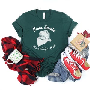 Könnte beinhalten: Ein grünes T-Shirt mit einer weißen Grafik von Weihnachtsmann und dem Text "Lieber Weihnachtsmann Bitte Definiere Gut". Das Shirt liegt auf einer weißen Oberfläche mit einem rot-schwarzen karierten Schal, einer weißen Tasse mit grünem Weihnachtsbaumdesign, blauen Jeans und roten Converse-Schuhen.