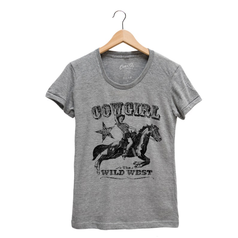 K&ouml;nnte beinhalten: Graues T-Shirt mit einem schwarz-wei&szlig;en Motiv einer Cowgirl, die ein Pferd reitet. Der Aufdruck lautet "Cowgirl" und "The Wild West".