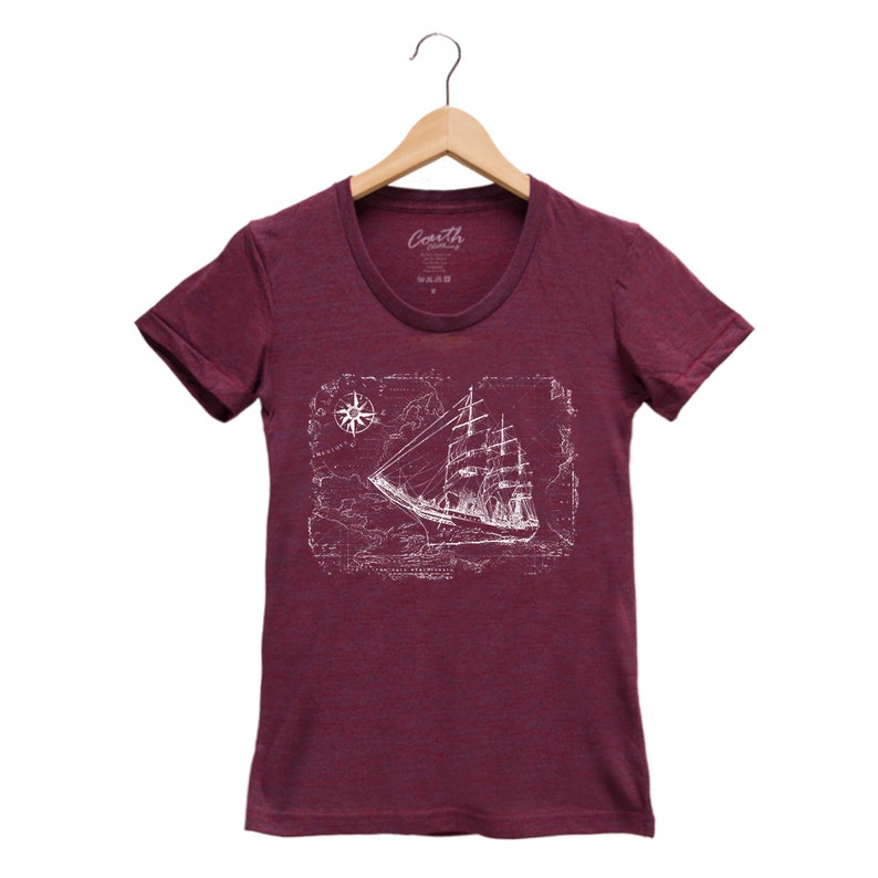 Puede incluir: Una camiseta color burdeos con un gr&aacute;fico blanco de un barco navegando en un mapa vintage con una rosa de los vientos.