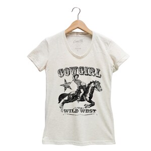 K&ouml;nnte beinhalten: Ein wei&szlig;es T-Shirt mit einem schwarz-wei&szlig;en Grafikdruck einer Cowgirl, die ein Pferd reitet. Die Grafik enth&auml;lt den Text "COWGIRL" und "The Wild West".