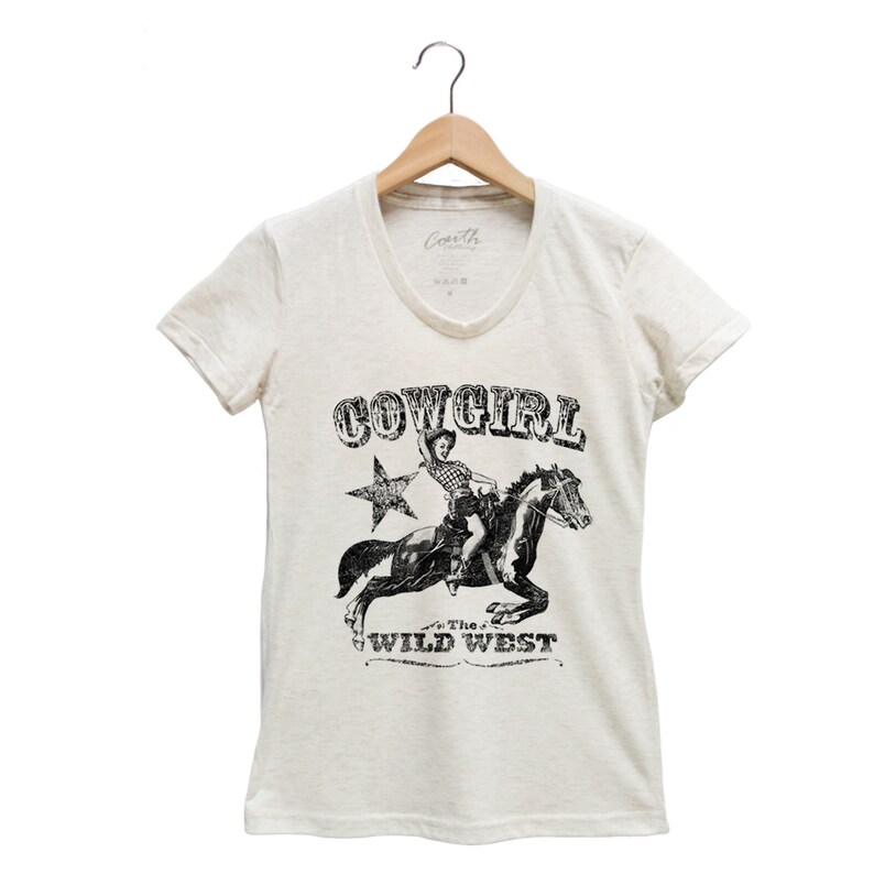 K&ouml;nnte beinhalten: Ein wei&szlig;es T-Shirt mit einem schwarz-wei&szlig;en Grafikdruck einer Cowgirl, die ein Pferd reitet. Die Grafik enth&auml;lt den Text "COWGIRL" und "The Wild West".