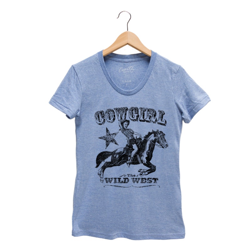 K&ouml;nnte beinhalten: Hellblaues T-Shirt mit einem Aufdruck einer Cowgirl, die ein Pferd reitet. Der Aufdruck ist in Schwarzwei&szlig; und enth&auml;lt den Text "COWGIRL" und "THE WILD WEST".