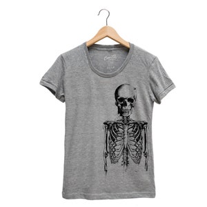 Puede incluir: Una camiseta gris jaspeada con una ilustraci&oacute;n en blanco y negro de un esqueleto en la parte delantera. El esqueleto est&aacute; de pie con los brazos a los lados. La camiseta tiene cuello redondo y mangas cortas.