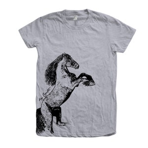 Op de afbeelding: Een heidekleurig t-shirt met een zwart-witte illustratie van een paard dat op zijn achterpoten staat. De illustratie is gesigneerd met "by Cavett".