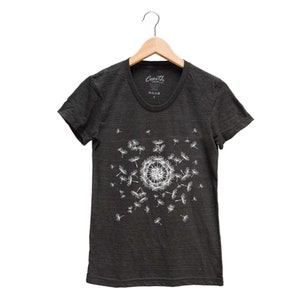 May include: A dark gray t-shirt with a white dandelion graphic. The dandelion is in the center of the shirt and has seeds blowing in the wind. The shirt has a crew neck and short sleeves. The text "Coath" is printed on the shirt.