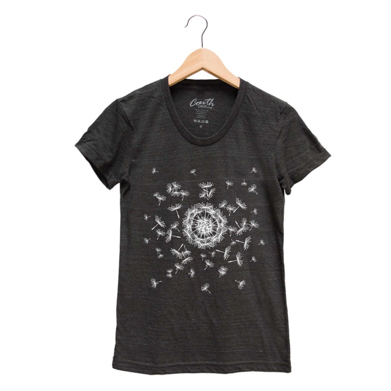 May include: A dark gray t-shirt with a white dandelion graphic. The dandelion is in the center of the shirt and has seeds blowing in the wind. The shirt has a crew neck and short sleeves. The text "Coath" is printed on the shirt.