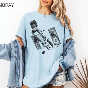Op de afbeelding: Een lichtblauw T-shirt met een zwart-witte illustratie van een satelliet. De tekst "By Costell" is onder de satelliet gedrukt.