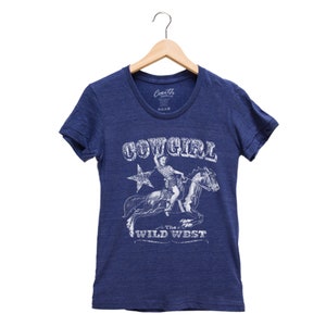 K&ouml;nnte beinhalten: Ein blaues T-Shirt mit einem Vintage-Grafikdruck einer Cowgirl, die ein Pferd reitet. Die Grafik enth&auml;lt den Text "COWGIRL" und "The Wild West".