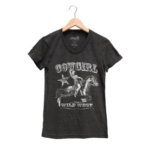 K&ouml;nnte beinhalten: Ein dunkelgraues T-Shirt mit einem wei&szlig;en Grafikdruck eines M&auml;dchens, das auf einem Pferd reitet. Die Grafik sagt "Cowgirl" und "The Wild West".