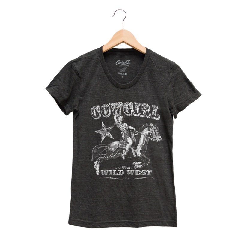 K&ouml;nnte beinhalten: Ein dunkelgraues T-Shirt mit einem wei&szlig;en Grafikdruck eines M&auml;dchens, das auf einem Pferd reitet. Die Grafik sagt "Cowgirl" und "The Wild West".