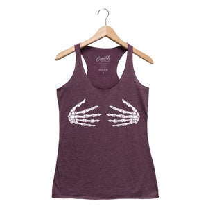 May include: A maroon racerback tank top with a white skeleton hand graphic. The tank top is hanging on a wooden hanger. The brand name "Coarth Clothing" is printed on the top of the tank top.