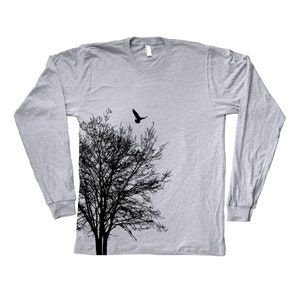 May include: A grey long-sleeved t-shirt featuring a black silhouette of a tree and a bird flying above it. The text "Coute" is printed on the bottom right corner of the shirt.