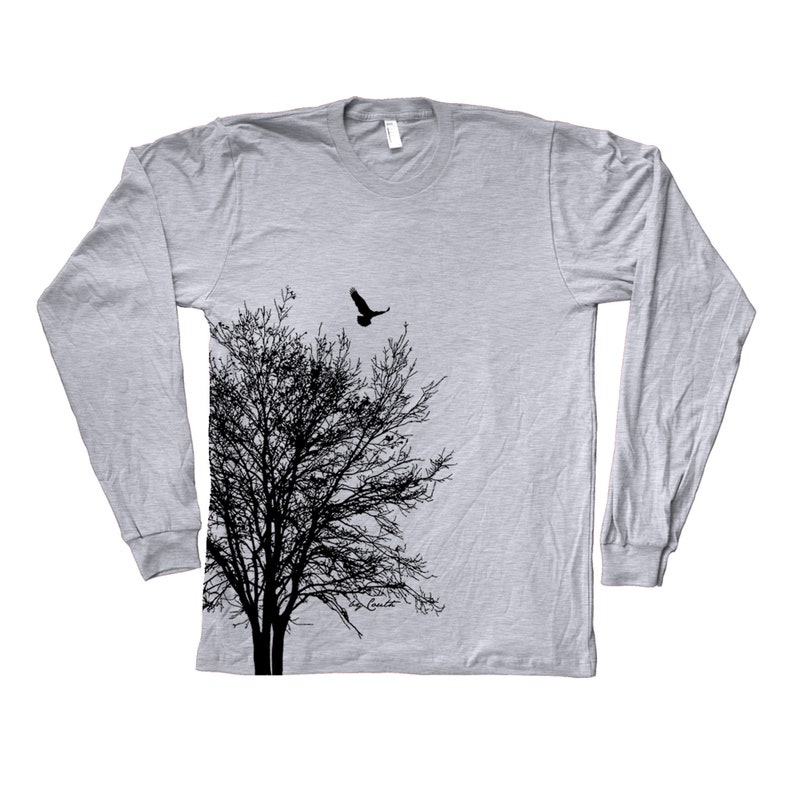 May include: A grey long-sleeved t-shirt featuring a black silhouette of a tree and a bird flying above it. The text "Coute" is printed on the bottom right corner of the shirt.