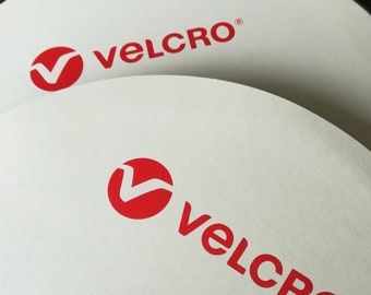 May include: Close-up of two white rolls of Velcro brand hook and loop tape. The rolls have a red logo with the word "VELCRO" printed in red.
