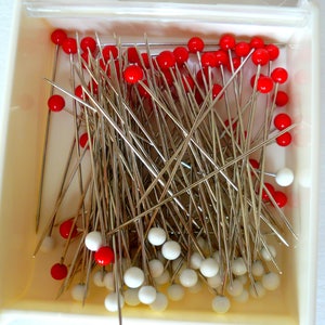 May include: A box filled with a large quantity of sewing pins with red and white plastic heads.