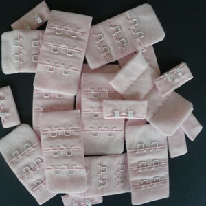 Puede incluir: Un montón de extensores de tirantes de sujetador de color rosa con ganchos de metal blanco. Los extensores están hechos de tela suave y tienen múltiples cierres de gancho y ojo.