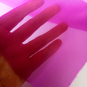 Peut inclure: Gros plan sur un tissu fin et transparent de couleur rose vif, &agrave; la texture en maille fine. Le tissu est tenu dans une main, ce qui met en &eacute;vidence sa d&eacute;licatesse et sa transparence.