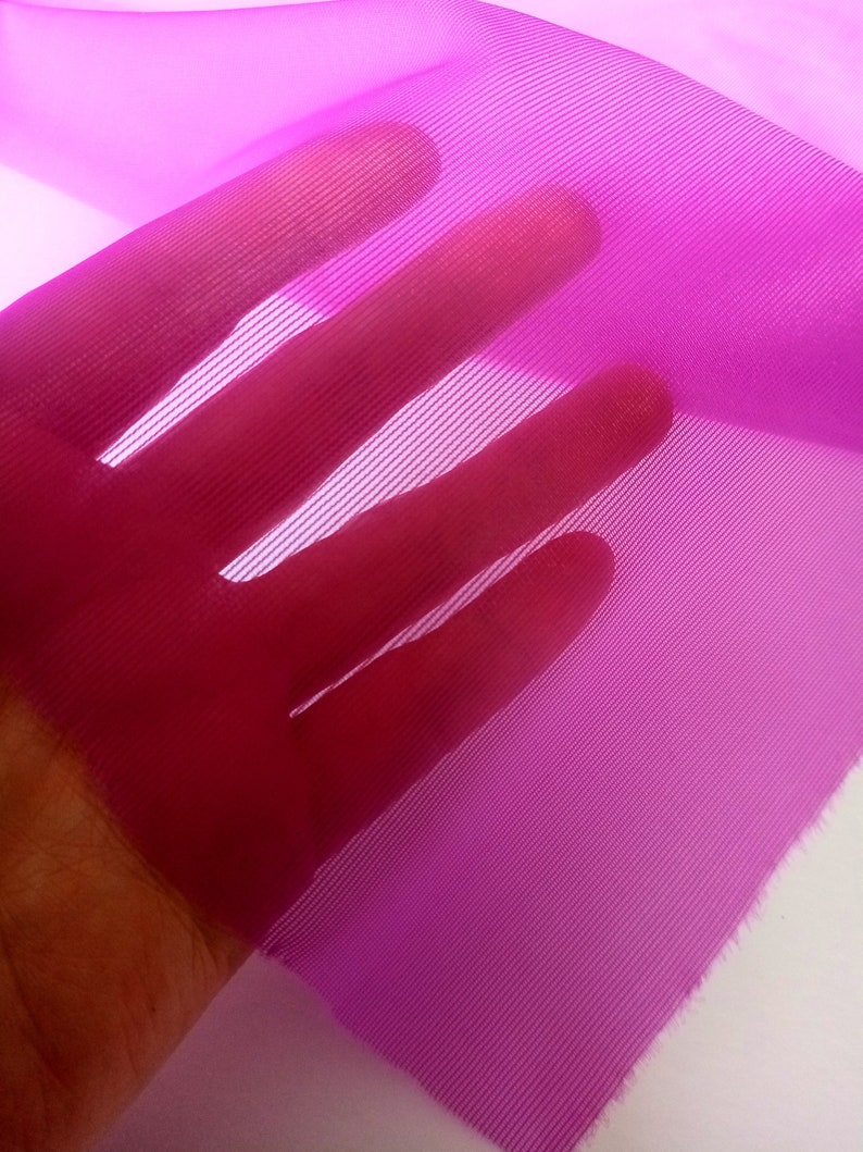 Peut inclure: Gros plan sur un tissu fin et transparent de couleur rose vif, &agrave; la texture en maille fine. Le tissu est tenu dans une main, ce qui met en &eacute;vidence sa d&eacute;licatesse et sa transparence.