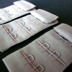 May include: Four beige bra strap extenders with metal hooks and eyes. The extenders are made of a soft, woven fabric.