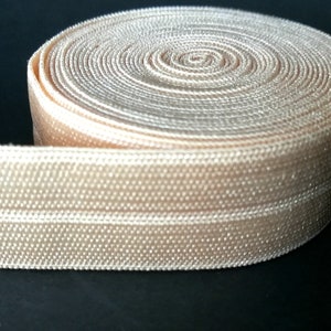 May include: A roll of beige elastic with a textured surface. The elastic is rolled up and has a white stripe running along the edge.