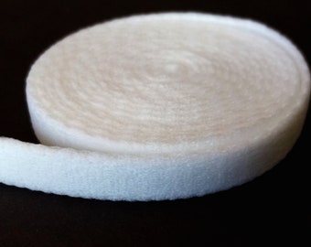Può includere: Un rotolo di tessuto bianco, morbido e strutturato, probabilmente feltro o un materiale simile, è mostrato su uno sfondo scuro. Il tessuto è arrotolato, rivelando la sua forma circolare e la consistenza del materiale.