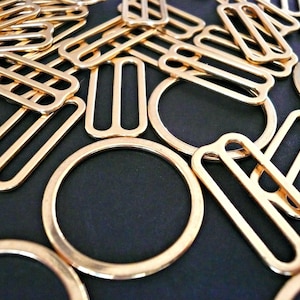 May include: A close-up of a group of gold-toned metal slider buckles. The buckles are arranged in a random pattern on a black surface.