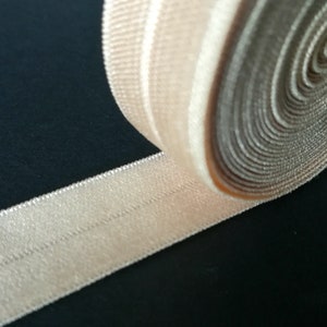 May include: A close-up of a beige elastic ribbon with a textured surface. The ribbon is rolled up and has a single line of stitching down the centre.