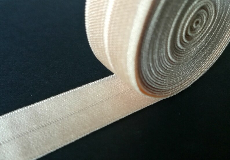 May include: A close-up of a beige elastic ribbon with a textured surface. The ribbon is rolled up and has a single line of stitching down the centre.