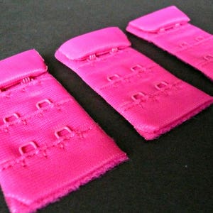 May include: Three pink bra strap extenders with metal hooks. Each extender has three rows of hooks.