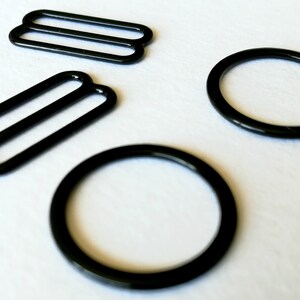 May include: Four black metal bra strap sliders. Two are round and two are rectangular with two parallel bars.