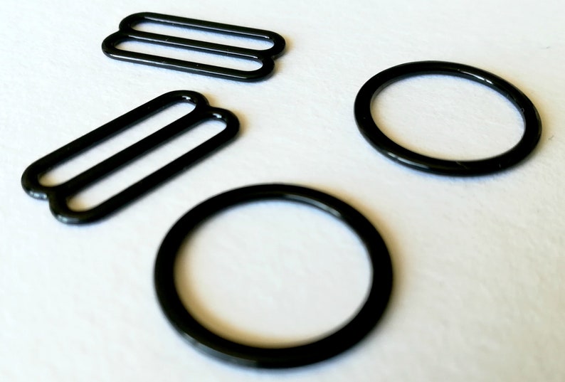 May include: Four black metal bra strap sliders. Two are round and two are rectangular with two parallel bars.