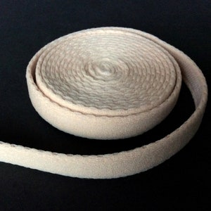 May include: A roll of beige fabric trim against a black background. The trim is tightly coiled, revealing its textured surface. The fabric appears to be a soft, neutral color, suitable for various sewing and crafting projects.