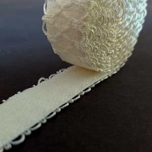May include: A roll of white elastic lace trim with a delicate floral pattern. The trim is rolled up and the edge of the lace is visible.