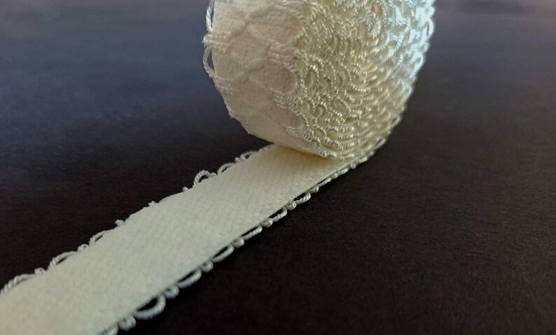 May include: A roll of white elastic lace trim with a delicate floral pattern. The trim is rolled up and the edge of the lace is visible.