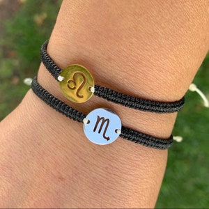 May include: Two black string bracelets with metal charms. The top charm is gold and features the astrological symbol for Leo. The bottom charm is silver and features the astrological symbol for Scorpio.