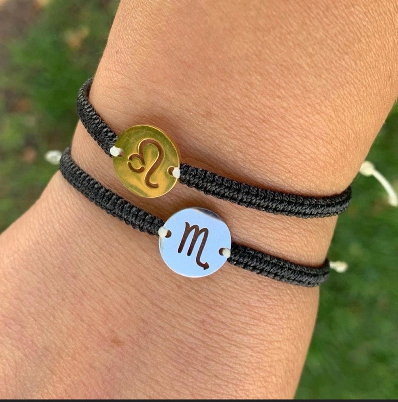 May include: Two black string bracelets with metal charms. The top charm is gold and features the astrological symbol for Leo. The bottom charm is silver and features the astrological symbol for Scorpio.