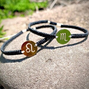 May include: Two black cord bracelets with silver and gold zodiac sign charms. The charms are round and feature the zodiac symbols for Leo and Scorpio.