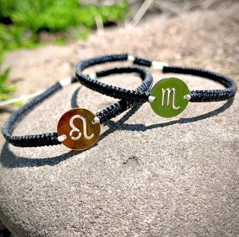 May include: Two black cord bracelets with silver and gold zodiac sign charms. The charms are round and feature the zodiac symbols for Leo and Scorpio.