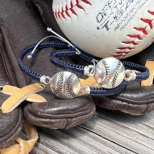 May include: Two navy blue braided bracelets with silver baseball charms. The bracelets are lying on a brown leather baseball glove.