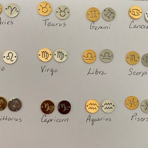 May include: Twelve gold and silver charms featuring the astrological signs of the zodiac. The signs are Aries, Taurus, Gemini, Cancer, Leo, Virgo, Libra, Scorpio, Sagittarius, Capricorn, Aquarius, and Pisces.