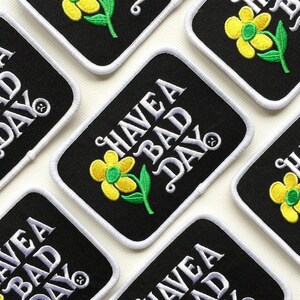 May include: A set of black iron-on patches with white text that reads "Have a Bad Day" and a yellow flower with green leaves.
